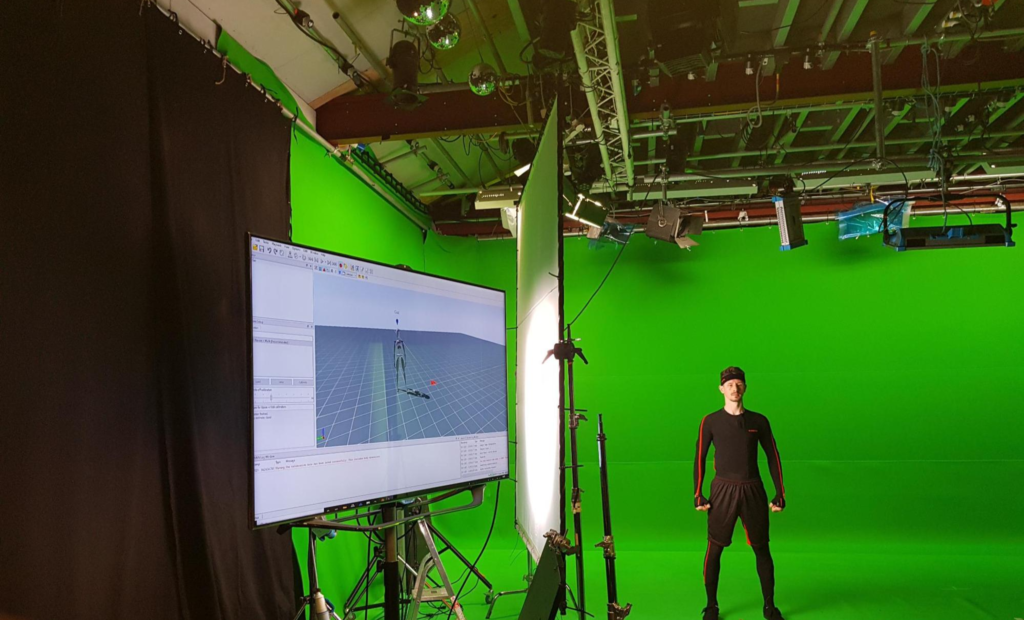 Motion capture performer in a green screen studio with tracking software displayed on a monitor.